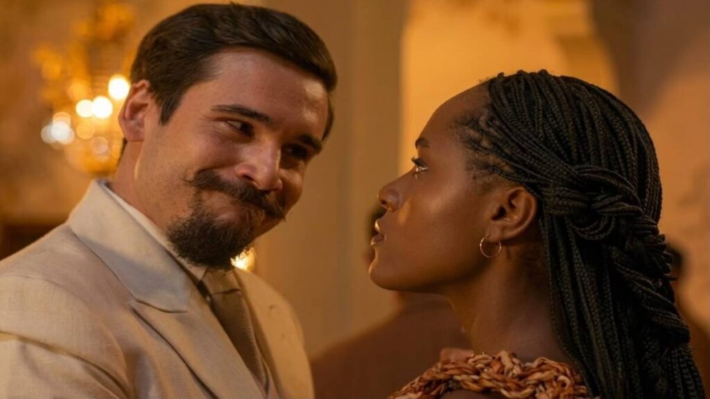 Mirinho, interpretado por Nicolas Prattes, observa Lúcia, vivida por Duda Santos, durante uma conversa em um salão iluminado em cena da novela A Nobreza do Amor.