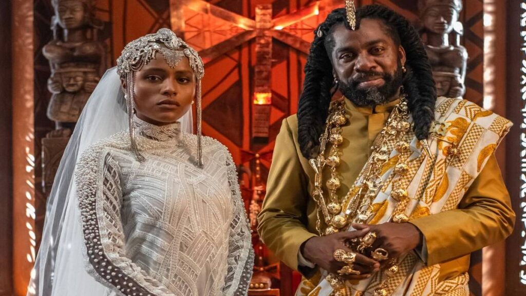 Os personagens Lúcia (Duda Santos) e Jendal (Lázaro Ramos) aparecem lado a lado em um cenário inspirado na cultura africana em A Nobreza do Amor. Ela veste um elaborado traje branco de cerimônia com véu e adornos prateados na cabeça, enquanto ele usa roupas douradas com colares e joias, em frente a um ambiente decorado com esculturas e elementos étnicos.