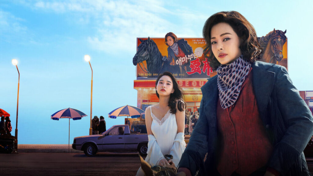 Pôster do dorama Madame Aema, da Netflix, mostra duas mulheres em destaque diante de um cinema na Coreia do Sul dos anos 1980, com um cartaz do filme fictício Aema ao fundo e cenário urbano retrô.
