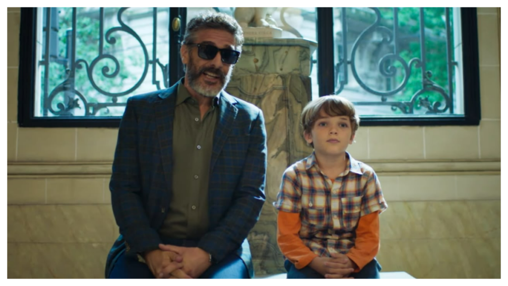 Cena do trailer de Vamos Consertar o Mundo mostra David Samarás, usando óculos escuros e blazer azul, sentado ao lado do menino Benito em um elegante salão iluminado por grandes janelas.