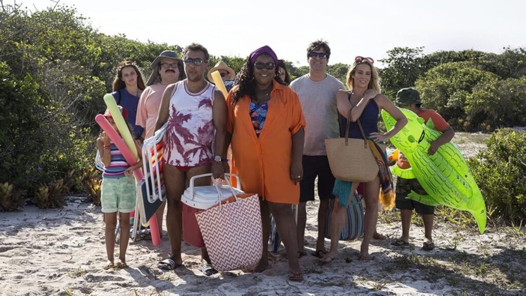Elenco de Os Farofeiros caminha por uma trilha de areia rumo à praia carregando cadeiras, caixas térmicas, boias e bolsas de viagem durante as férias.